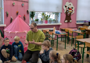 Mężczyzna z dziećmi siedzi na dywanie i objaśnia reguły gry